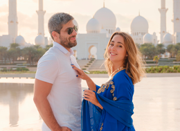 Um casal sorridente posa em frente à grandiosa e luminosa arquitetura branca de uma mesquita com minaretes esguios e reflexos na água. A mulher veste um elegante vestido azul com detalhes dourados e um véu, enquanto o homem veste uma camisa polo branca e óculos de sol. A atmosfera é de um dia ensolarado de celebração ou turismo em um local icônico.